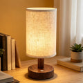Cordless rechargeable table lamp with linen shade and wood grain base glowing warmly on a bedside shelf
